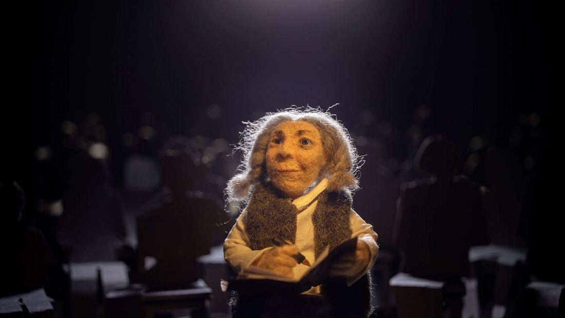 Mama Micra - Still - An elderly woman made of felt writing in her notebook.