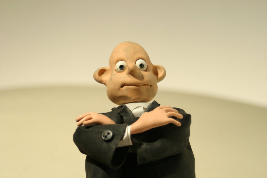 Closed - Still - A man made of clay looking down at the camera with his arms crossed.